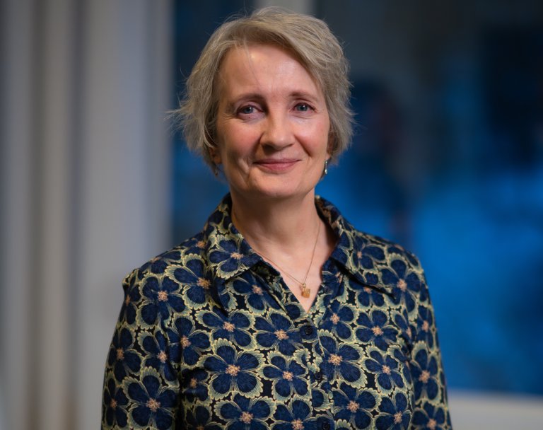 Foto av en kvinne. Hun har mellomblondt hår, en blomstrete bluse og har på seg et gullsmykke. Hun smiler til kamera.