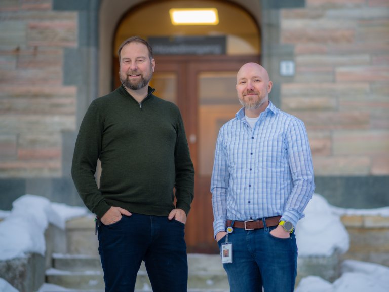 To menn fotografert utenfor en steinbygning. Begge ser i kameraet og smiler.