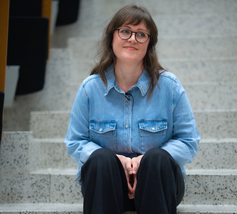 Foto av en kvinne som sitter i en trapp. Hun har mørkt hår og briller.